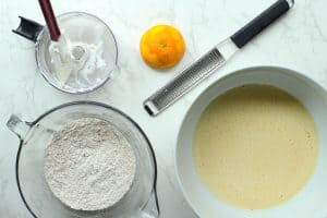 A food processor with blended cottage cheese residue, a halved orange next to hydroplane, separate bowls of dry ingredients and wet.