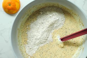 Orange poppyseed dry ingredients being incorporated and folded into the wet ingredients with a halved orange in the corner.