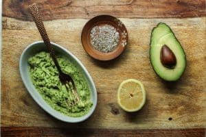 Mashed avocado with a fork in a white bowl, a half lemon, a halved avocado with the pit, and a small dish with sea salt and pepper.