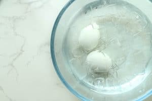 Two cooked eggs in an ice bath in a clear bowl on a white granite surface.