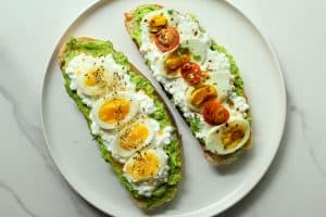 Avocado cottage cheese toast on a white plate and a white granite surface. One with chili oil and honey and one with tomatoes and balsamic.