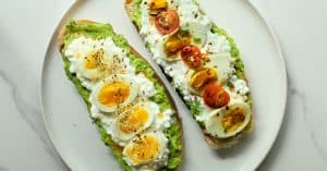 Avocado cottage cheese toast on a white plate and a white granite surface. One with chili oil and honey and one with tomatoes and balsamic.
