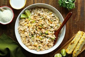 Mexican street corn pasta salad in a white bowl with lime wedges, a bowl of cotija, corn cobs, green napkin on wooden surface.