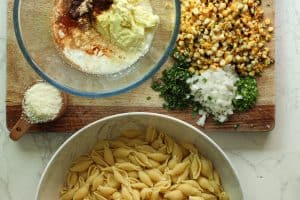 Mexican street corn pasta salad ingredients with pasta in one bowl, dressing ingredients in another, and chopped veggies and corn.