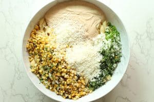 Mexican street corn pasta salad ingredients like chopped veggies, charred corn, chipotle dressing, and cotija on a white granite surface.