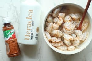 A bowl of shrimp sprinkled with tajin in a white bowl and a bottle of Heraclea extra-virgin olive oil and a bottle of tajin.