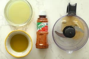 Fresh lime juice in a dish, olive oil in a yellow rimmed dish, a food processor with honey and garlic, and a bottle of tajin in the center.