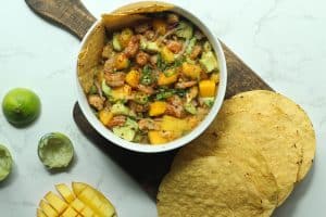 A bowl of shrimp, avocado, and mango salad with tostadas on the side and already squeezed lime halves.