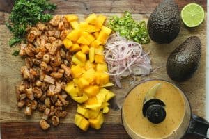 Shrimp, mango, red onion, and cilantro cut on a wooden cutting board with two whole avocados, a halved lime, and chili-lime vinaigrette.