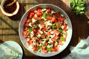 Watermelon salad with feta cheese in a white bowl on a wooden surface with a bowl of chili crisp, fresh herbs and a white napkin.