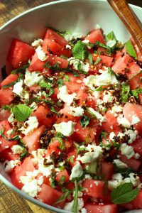 Watermelon feta salad sprinkled with homemade chili crisp and fresh herbs such as mint and basil in a white bowl on wooden surface.