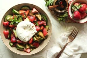 A bowl of cucumber strawberry salad topped with a cut open burrata, a cup of balsamic glaze, and a bowl of strawberries.