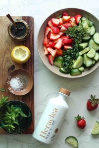 A bowl of strawberries, cucumbers, mint, basil, a bottle of Heraclea, balsamic glaze, a halved lemon for cucumber strawberry salad.