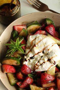 Close up of cucumber strawberry salad in a bowl with an open burrata cheese ball drizzled with balsamic glaze and a mint sprig.