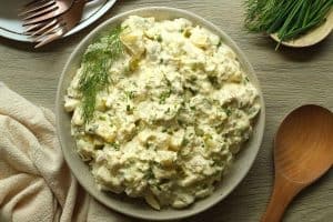Potato salad with dill pickles and dill sprig in a beige speckled bowl with neutral tones in the napkin, wooden surface, and spoon.