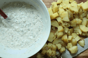 Potato salad dressing ingredients combined in a white bowl next to diced potatoes with a knife on a cutting board.