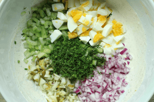 Potato salad with dill pickles dressing in a white bowl along with diced onion, pickles, celery, hard-boiled eggs, chives, and dill.