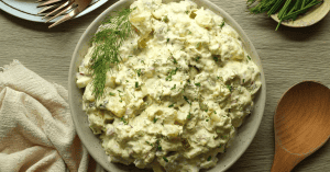 Potato salad with dill pickles and dill sprig in a beige speckled bowl with neutral tones in the napkin, wooden surface, and spoon.