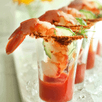 Cocktail party food close up of shrimp cocktail shooters with a slice of cucumber and sprig of dill on a white plate with ice.