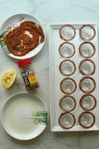 A white rectangular plate with shot glasses rimmed with old bay, a bottle of the seasoning, a plate with lemon juice, and halved lemon.