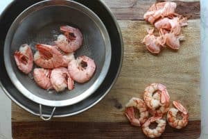 Cooked shrimp draining in a fine mesh strainer and 5 shrimp on the side on a cutting board that have already been peeled.