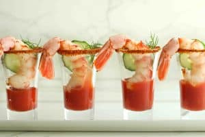 4 shrimp cocktail shooters lined in a row in shot glasses with a single shrimp hanging over the edge and a slice of cucumber.