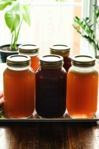 A bright scene with natural light shining through jars of homemade bone broth making the golden color pop with plants in the background.