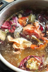 Roasted veggie scraps and a chicken carcass in an Instant Pot filled with water with oil or fat droplets on the surface.