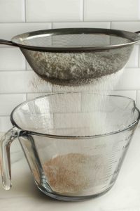 Flour, spices, baking powder, and baking soda being sifted through a find mesh strainer into a glass bowl.
