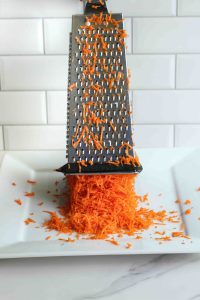 A box grater being lifted with shredded carrots on it and shredded carrots falling out from the bottom on a white plate.