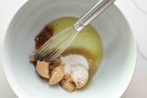 Brown sugar, white sugar, and avocado oil in a white bowl with a whisk before being whisked.