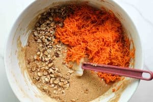 Carrot cake loaf cake batter in a white bowl with chopped walnuts and shredded carrots before being mixed in with a rubber spatula.