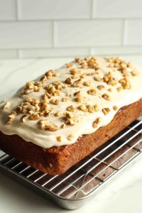A golden-brown carrot loaf cake frosted with cream cheese cottage cheese frosting and topped with walnuts on a wire rack.