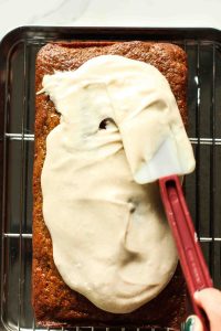 Frosting being spread on a carrot loaf cake with a rubber spatula.