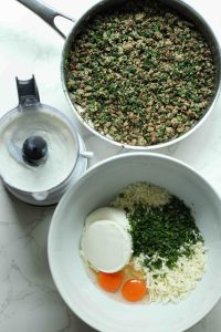 Ground beef and spinach cooked in a pan, a bowl of eggs, cheese, and herbs, and a food processor with blended cottage cheese.