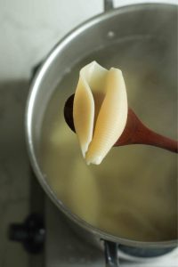 A cooked jumbo pasta shell on a wooden spoon over a pot of boiling water with more pasta.