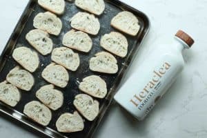 Crostini bread slices line on a silver sheet baking pan with a white bottle of Heraclea olive oil on the side.