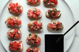 Bruschetta on crostini with goat cheese on a round white plate with a square white dish with balsamic glaze.