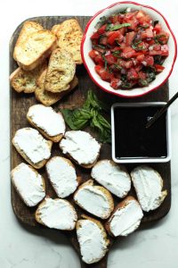 Crostini with goat cheese and plain crostinis displayed on a wooden cutting board with a dish of tomato bruschetta and balsamic glaze.