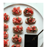 How to make bruschetta: pieces of crostini topped with goat cheese and bruschetta on a white plate with a dish of balsamic glaze.