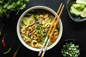 Tom yum noodle soup in a white bowl with brown and white chopsticks, lime wedges and scallions in separate bowls, and thai chilis.