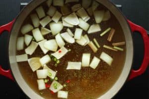 A dutch oven filled with chicken bone broth, onion chunks, sliced thai chiles, ginger, and lemongrass.