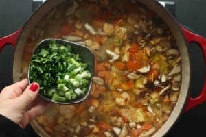 A bowl of sliced scallions and cilantro being held over the pot of tom yum noodle soup before being poured in.